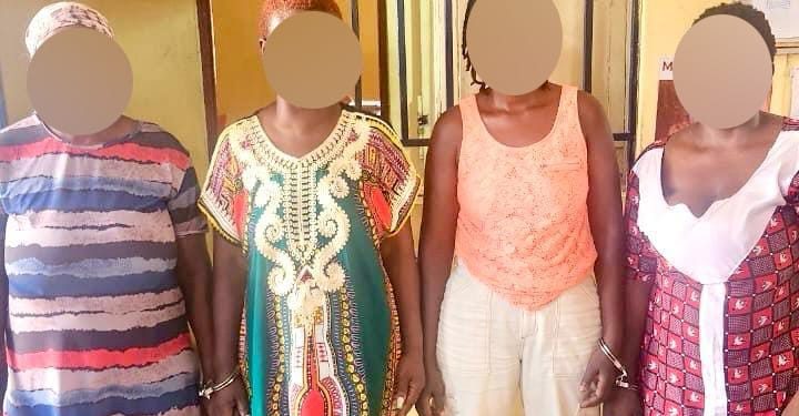 Four people stand side by side indoors with faces blurred; they wear colorful casual outfits (striped shirt, green patterned tunic, orange sleeveless top, and pink patterned dress). The warm yellow walls suggest a casual group photo setting.