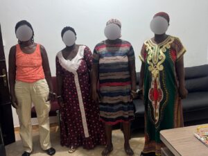 Four women with blurred faces pose indoors against a light wall, each wearing a distinct colorful dress.