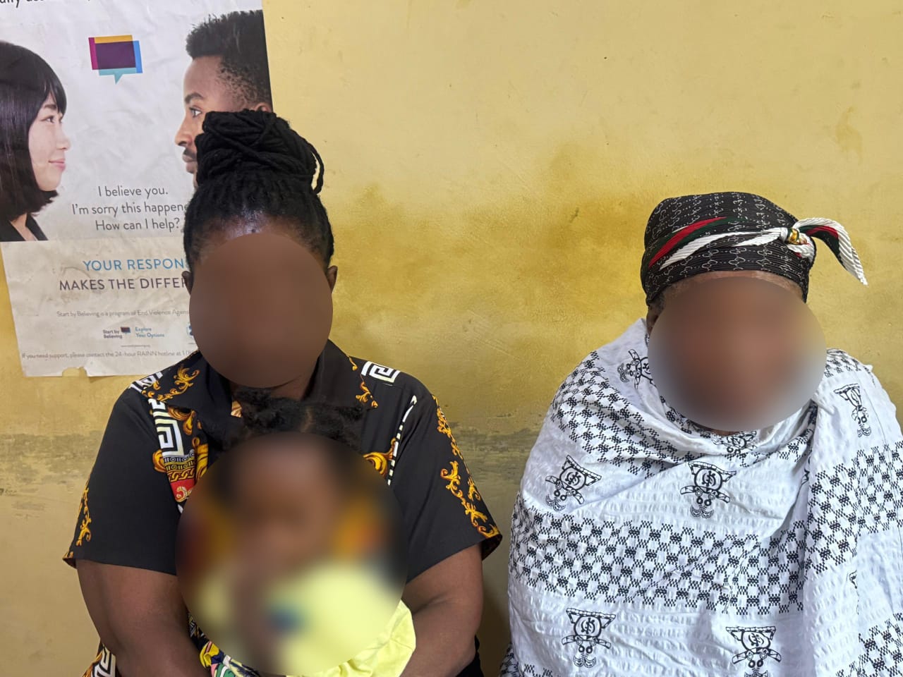 Two women sit against a yellow wall; the left woman holds a blurred child in yellow, the right woman wears a white patterned shawl and head wrap.