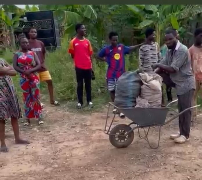 Group of villagers standing on a dirt path with a wheelbarrow loaded with bags, in a rural garden setting Hot sunny day with greenery in background.