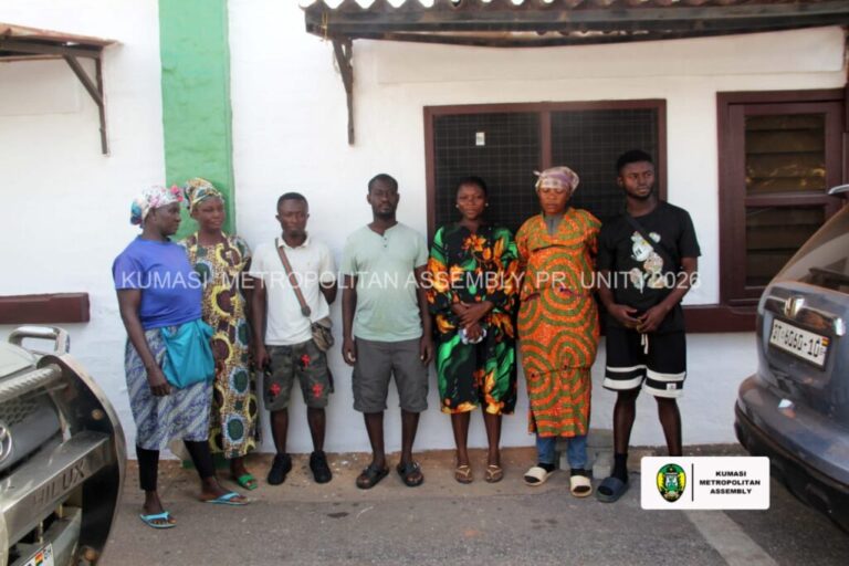 Eight people stand in a line outside a white building with a green pillar, wearing colorful traditional clothing; two cars are parked nearby.