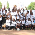 Group of diverse youths and coaches in white shirts on a dirt soccer field, hands raised in celebration around a central soccer ball.