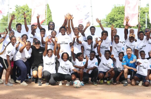 Group of diverse youths and coaches in white shirts on a dirt soccer field, hands raised in celebration around a central soccer ball.