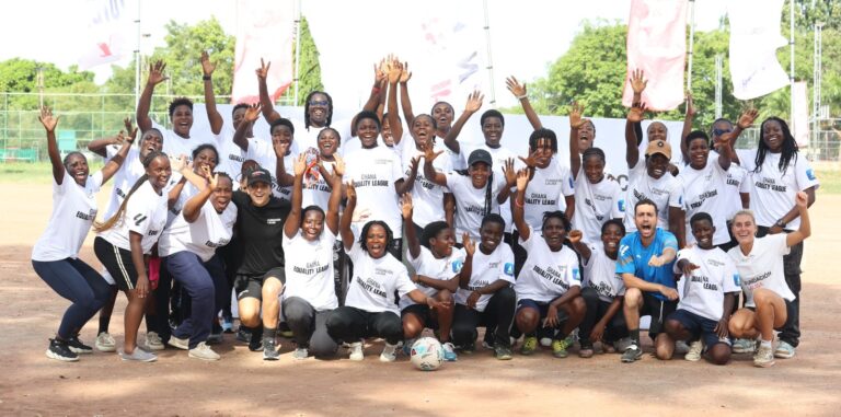 Group of diverse youths and coaches in white shirts on a dirt soccer field, hands raised in celebration around a central soccer ball.