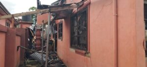 Side of a pink stucco building with broken windows, charred beams, and a collapsed roof supported by wooden posts; debris on the ground.