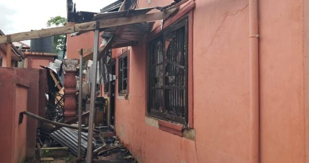 Side of a pink stucco building with broken windows, charred beams, and a collapsed roof supported by wooden posts; debris on the ground.