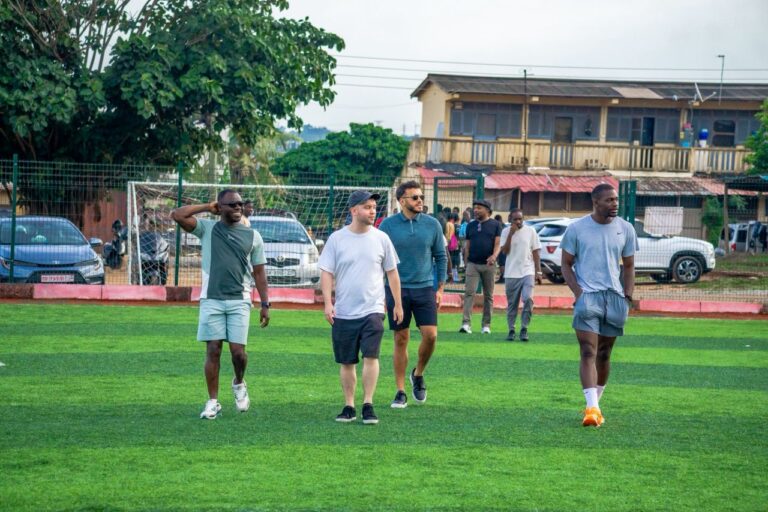 Group of men walking on a green artificial soccer field, with a goal and fence in the background and spectators behind them.