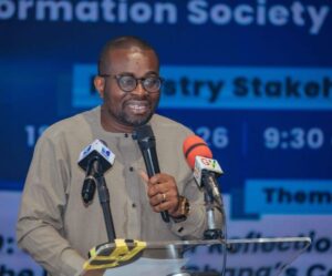 Man wearing glasses and a beige traditional outfit speaks into a handheld microphone at a podium with two microphones, at a conference stage.