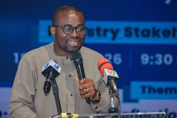 WhatsApp-Image-2026-05-19-at-171935 | Ghanaguardian.com Man wearing glasses and a beige traditional outfit speaks into a handheld microphone at a podium with two microphones, at a conference stage.