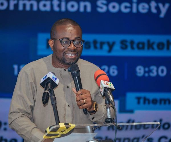Man wearing glasses and a beige traditional outfit speaks into a handheld microphone at a podium with two microphones, at a conference stage.