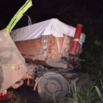 Night scene of a damaged dump truck off the road, with a tarp-covered load and a red cylindrical tank visible, surrounded by tall grass at night.