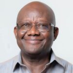 Portrait of a smiling bald man wearing glasses in a light striped shirt against a pale background.