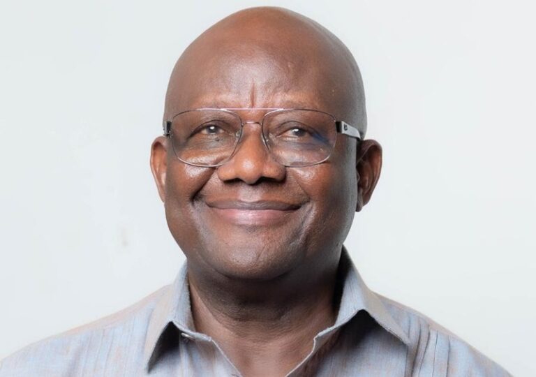 Portrait of a smiling bald man wearing glasses in a light striped shirt against a pale background.