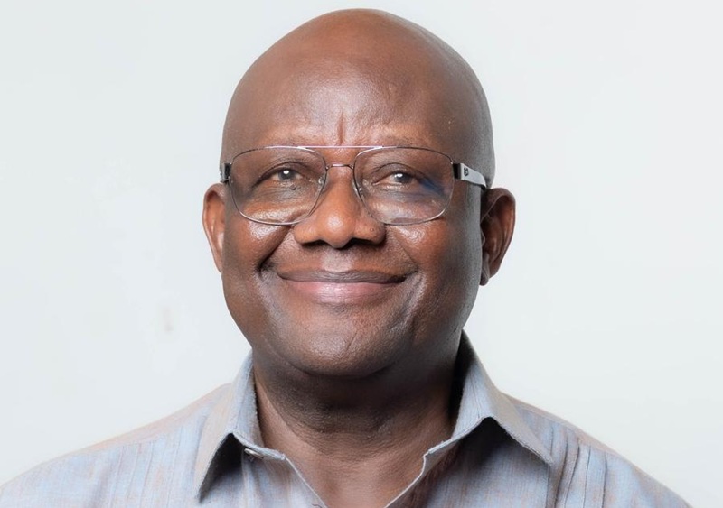Portrait of a smiling bald man wearing glasses in a light striped shirt against a pale background.