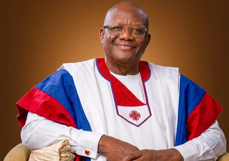 Smiling older man in white ceremonial attire with red and blue accents, seated against a brown gradient backdrop.