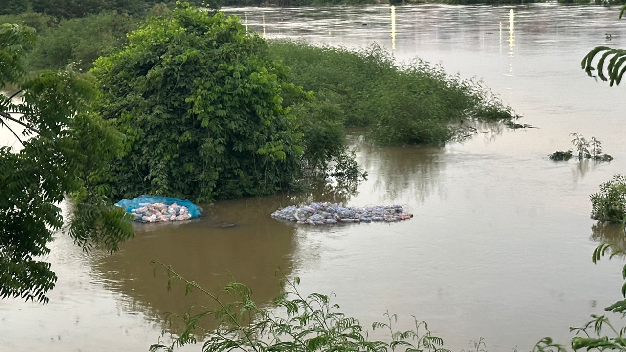 WhatsApp-Image-2026-05-28-at-074502-1 | Ghanaguardian.com Flooded riverside with piles of bags on a blue tarp near partially submerged bushes and trees in muddy water.