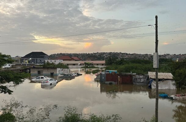 WhatsApp-Image-2026-05-28-at-074503-1 | Ghanaguardian.com Flooded residential scene at sunset: cars partially submerged in muddy water, with makeshift homes along the bank and a hillside in the distance.