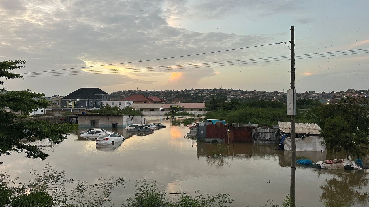WhatsApp-Image-2026-05-28-at-074503-1 | Ghanaguardian.com Flooded residential scene at sunset: cars partially submerged in muddy water, with makeshift homes along the bank and a hillside in the distance.