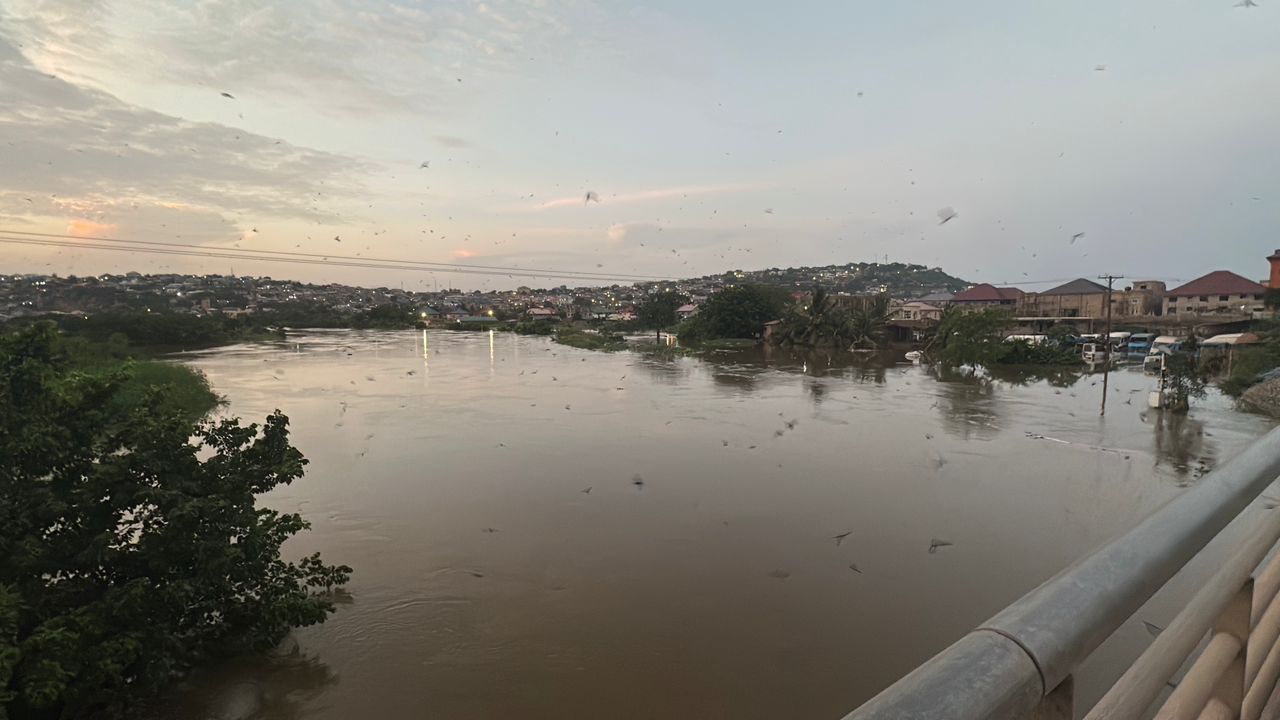 WhatsApp-Image-2026-05-28-at-074503 | Ghanaguardian.com River overflowing into a town, viewed from a bridge at dusk with muddy water and flooded streets in the distance.