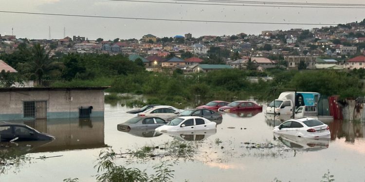 WhatsApp-Image-2026-05-28-at-074504-750x375 | Ghanaguardian.com Cars submerged in floodwater along a street, with a residential area and hills in the background
