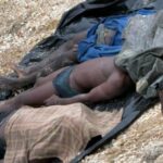 Several people lie prone on a black tarp spread on rocky ground, with torn clothing; a man sits nearby, suggesting casualties or injured individuals.