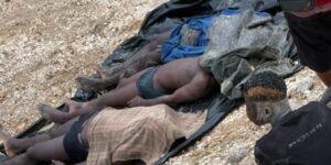 Several people lie prone on a black tarp spread on rocky ground, with torn clothing; a man sits nearby, suggesting casualties or injured individuals.