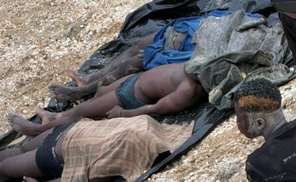 Several people lie prone on a black tarp spread on rocky ground, with torn clothing; a man sits nearby, suggesting casualties or injured individuals.
