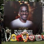 Outdoor memorial banner showing a large smiling portrait of Dr. Yaw Odoomfo, with floral wreaths in front and a metal stage structure nearby.