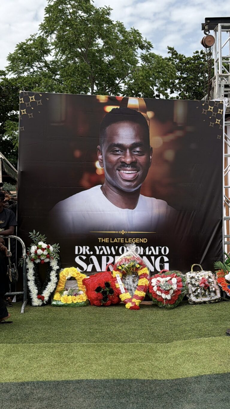 Outdoor memorial banner showing a large smiling portrait of Dr. Yaw Odoomfo, with floral wreaths in front and a metal stage structure nearby.