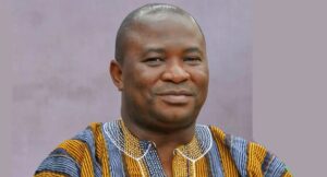 Portrait of a smiling man wearing a colorful striped traditional African shirt against a muted purple background.