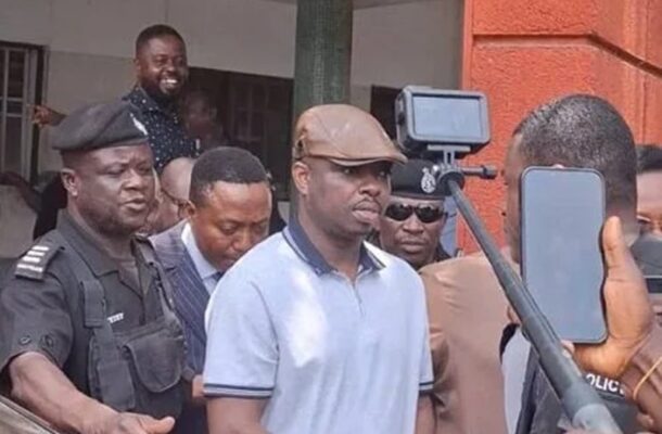 Man in a light blue polo and brown cap is escorted by police as onlookers film with smartphones.