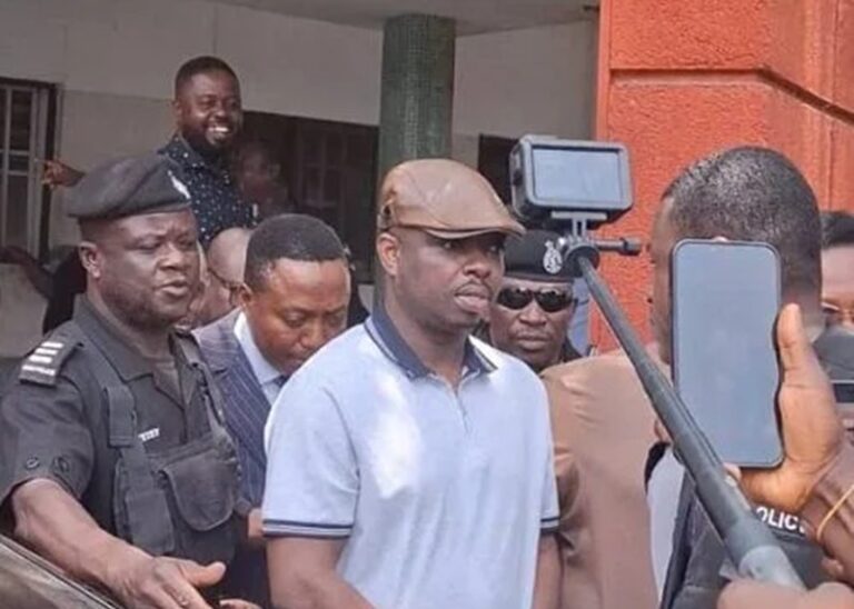 Man in a light blue polo and brown cap is escorted by police as onlookers film with smartphones.