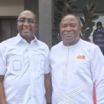 Two men in light-colored outfits smile for the camera at an outdoor event.