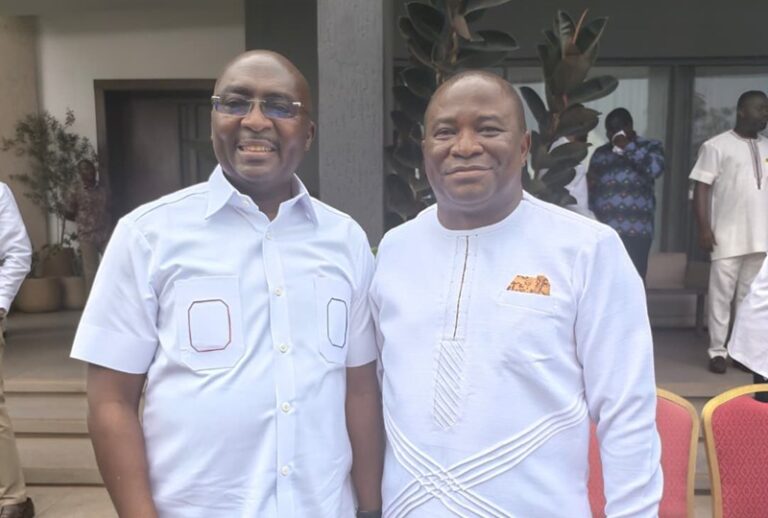 Two men in light-colored outfits smile for the camera at an outdoor event.