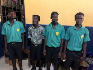 Four male students in green uniforms with crest badges stand indoors; one wears a patterned shirt and all faces are pixelated for privacy.