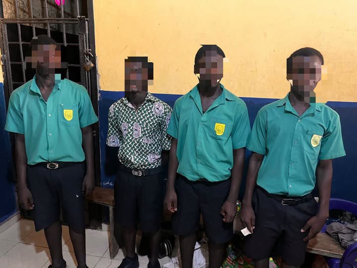 Four male students in green uniforms with crest badges stand indoors; one wears a patterned shirt and all faces are pixelated for privacy.