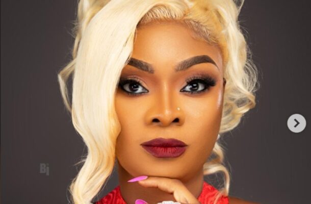 afaglo | Ghanaguardian.com Portrait of a person with platinum blonde hair, bold makeup, red lips, and pink nails posing with a hand near the chin against a dark background.