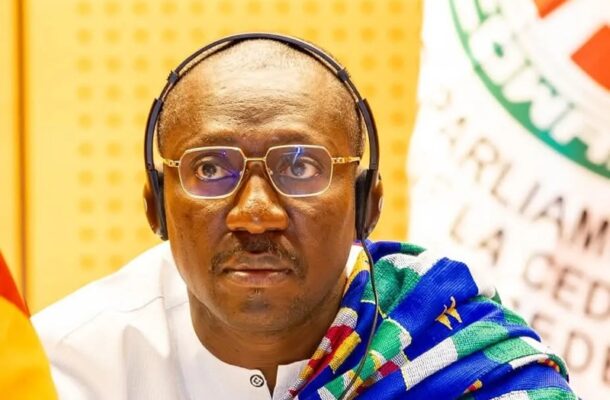 Portrait of a man wearing over-ear headphones and rectangular glasses, dressed in white with a colorful scarf, at a conference setting.
