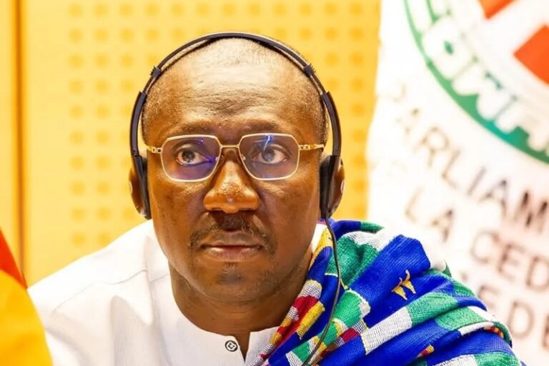 Portrait of a man wearing over-ear headphones and rectangular glasses, dressed in white with a colorful scarf, at a conference setting.