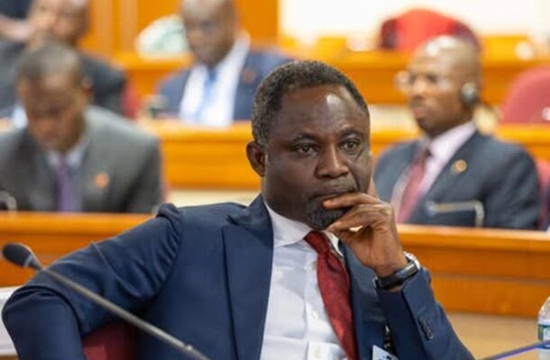 African man in a blue suit and red tie, seated at a conference table with hand on chin, thoughtful expression.