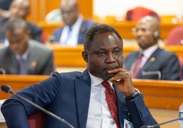 African man in a blue suit and red tie, seated at a conference table with hand on chin, thoughtful expression.