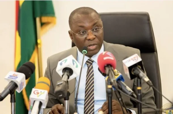 A man in a gray suit and striped tie speaks into a microphone at a formal press conference, with multiple news microphones in front and a flag in the background.