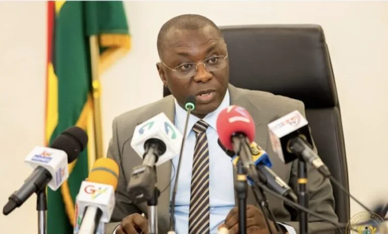 A man in a gray suit and striped tie speaks into a microphone at a formal press conference, with multiple news microphones in front and a flag in the background.
