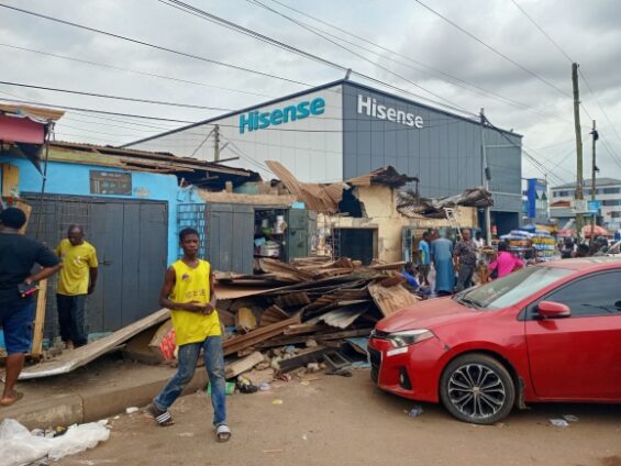 Collapsed wooden structure in front of a Hisense storefront, with bystanders and a red car nearby.