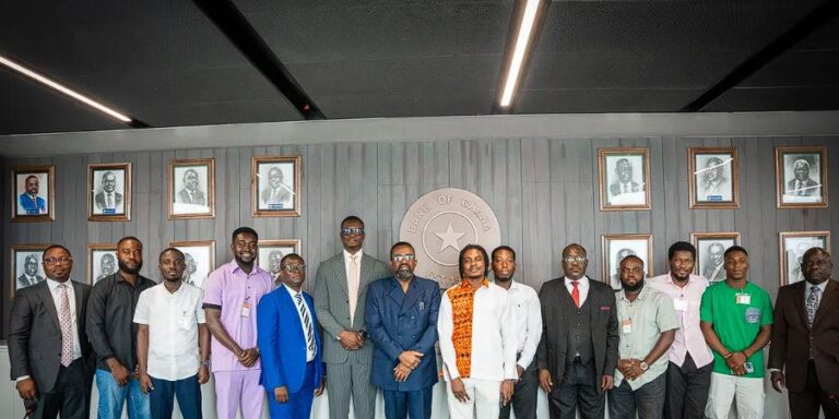 Group of professionally dressed people posing for a formal photo in front of a government seal and framed portraits on the wall.