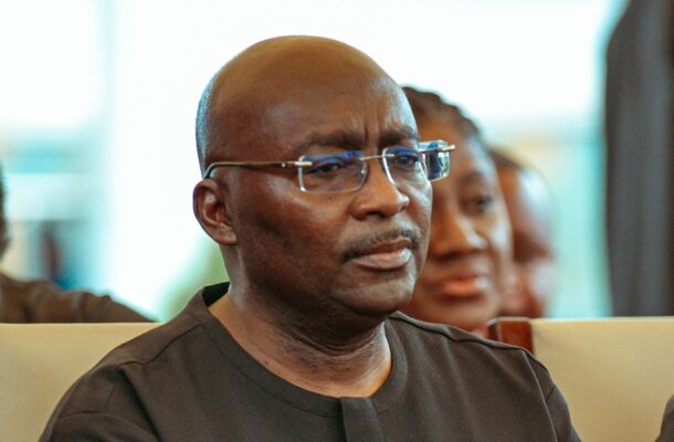bawumia | Ghanaguardian.com Man wearing glasses and a black shirt listening intently at an indoor event or conference venue, seated in the audience.