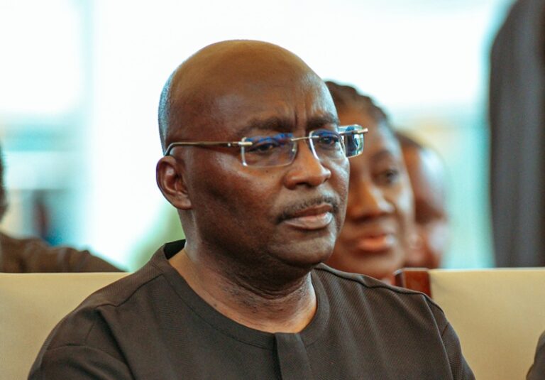 Man wearing glasses and a black shirt listening intently at an indoor event or conference venue, seated in the audience.