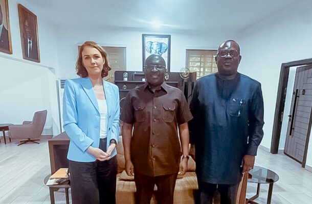 Three people posing for a photo in an office lounge: a woman in a blue blazer, and two men in brown and dark traditional outfits.