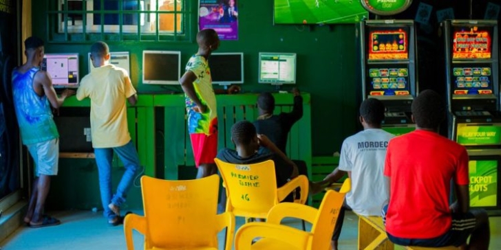 People in a green-lit arcade gathered around computer stations, with yellow chairs in the foreground and game monitors in use.