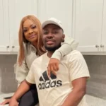 A woman and man lean on a kitchen counter, smiling at the camera, with white cabinets in the background. The man wears an Adidas shirt and a cap; the woman rests her arm on his shoulder.